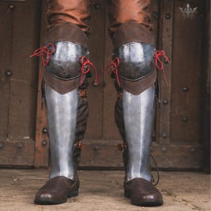 May include: A pair of leg armour pieces featuring polished steel knee cops and greaves attached to brown leather boots. The knee armour is secured with bright red laces, while the lower leg plates are fastened with brown leather straps and buckles.