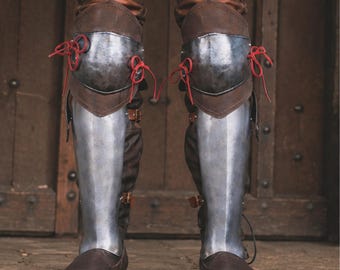 May include: A pair of leg armour pieces featuring polished steel knee cops and greaves attached to brown leather boots. The knee armour is secured with bright red laces, while the lower leg plates are fastened with brown leather straps and buckles.