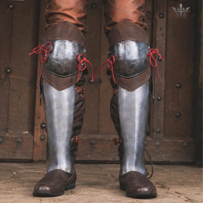May include: A pair of leg armour pieces featuring polished steel knee cops and greaves attached to brown leather boots. The knee armour is secured with bright red laces, while the lower leg plates are fastened with brown leather straps and buckles.