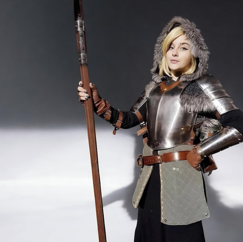 May include: A person in a medieval costume, featuring a metal breastplate, fur-trimmed hood, and brown leather gloves, holding a long wooden spear. The outfit includes a brown belt and a quilted skirt. The background is a neutral grey.