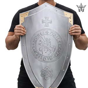 May include: A silver shield with a cross and a circular emblem depicting a knight on horseback. It features gold corner accents and rivet detailing. The shield has a medieval design.
