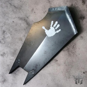 Op de afbeelding: Een metalen schild met een handafdrukontwerp. Het schild heeft een doffe zilveren kleur met een witte handafdruk in de rechterbovenhoek. Het schild heeft twee puntige randen aan de onderkant en is vastgeniet.