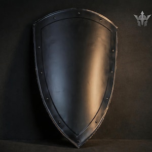 Puede incluir: Un escudo negro con la parte superior redondeada y la inferior puntiaguda. El escudo tiene un acabado oscuro y mate con remaches a lo largo de los bordes. Un logotipo de cresta es visible en la esquina superior derecha. El escudo es probablemente para exhibición o disfraz.