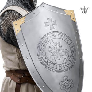 May include: A silver-colored medieval shield with a cross emblem and a circular design featuring two knights on horseback. The shield has gold-colored corner accents and rivets along the edges.