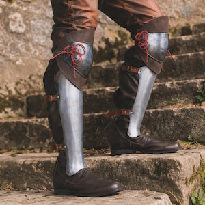 May include: Dark brown leather boots featuring integrated steel greaves and knee plates. The metal armour components are secured with brown leather straps and buckles, while the knee sections are fastened with thin red cord lacing.
