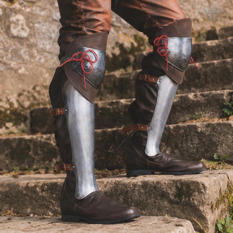May include: Dark brown leather boots featuring integrated steel greaves and knee plates. The metal armour components are secured with brown leather straps and buckles, while the knee sections are fastened with thin red cord lacing.