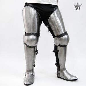 May include: A pair of polished steel leg armor pieces covering the thighs, knees, and shins, secured with black leather straps and buckles. The metal plates feature rounded knee caps and pointed foot guards, worn over black trousers.