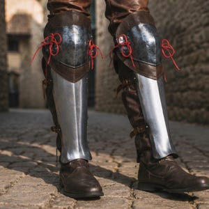 May include: A pair of metal greaves and knee armour worn over brown leather boots and trousers. The silver-toned steel leg guards feature polished surfaces and are secured with bright red laces at the knees and brown leather straps along the calves.