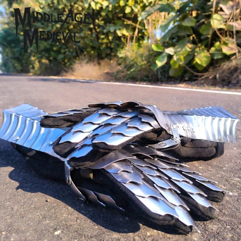 May include: A pair of medieval gauntlets made of shiny, silver-colored metal plates, with a black fabric interior. The gauntlets are lying on a paved surface, with the text "MIDDLE AGE MEDIEVAL" visible in the background.