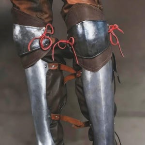 May include: A pair of leg guards featuring polished steel plates covering the shins and knees, layered over brown leather material. The metal components are secured with brown leather straps and buckles, while bright red leather laces tie at the knees.