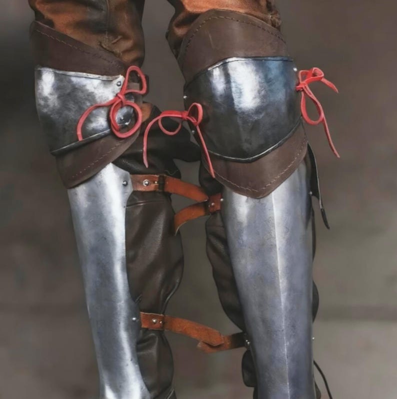 May include: A pair of leg guards featuring polished steel plates covering the shins and knees, layered over brown leather material. The metal components are secured with brown leather straps and buckles, while bright red leather laces tie at the knees.