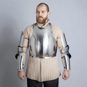 May include: A shiny, silver breastplate and arm armour. The armour is worn over a quilted, beige-coloured garment. The person is standing against a light grey background. The armour appears to be medieval or historical in style.