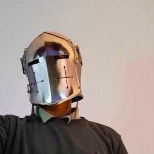 May include: A polished silver knight's helmet with a cross design on the front. The helmet has a reflective surface and is adorned with decorative chains. The image shows the helmet from the chest up, against a neutral background.