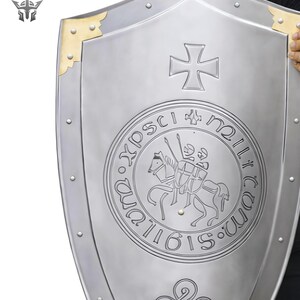 May include: A silver-colored shield with a cross emblem and a circular inscription featuring a knight on horseback. The shield has gold-colored corner accents and is held by a person.