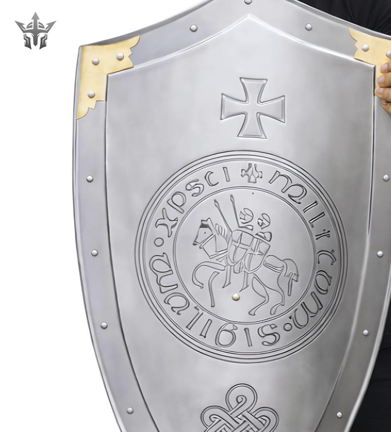 May include: A silver-colored shield with a cross emblem and a circular inscription featuring a knight on horseback. The shield has gold-colored corner accents and is held by a person.