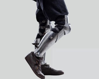 May include: Polished metal leg armour, comprising articulated knee and shin plates, fastened with leather straps and buckles. The armour is worn over dark fabric, and paired with brown leather boots. The design suggests historical reenactment or fantasy-themed clothing.