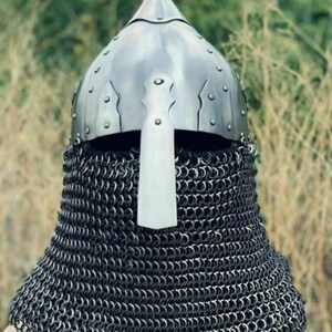 May include: A medieval-style helmet with a pointed top and a face guard. The helmet is made of shiny metal and is riveted. A chainmail coif extends down to the neck. The background is blurred with green and brown tones.