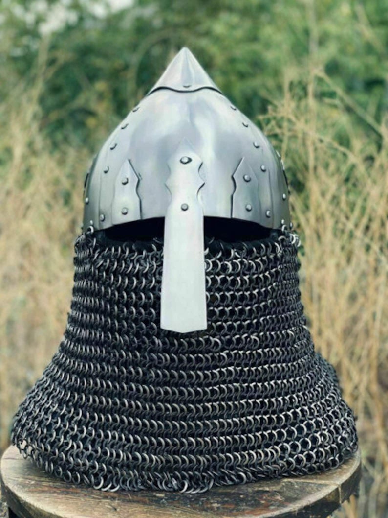 May include: A medieval-style helmet with a pointed top and a face guard. The helmet is made of shiny metal and is riveted. A chainmail coif extends down to the neck. The background is blurred with green and brown tones.