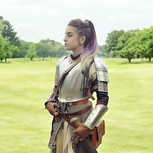 May include: A person wearing silver armour, brown leather gloves, and holding a sword. The armour covers the chest, shoulders, and arms. The person is standing in a grassy field with trees in the background.