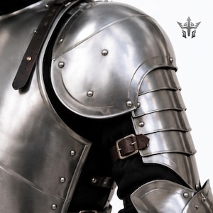 May include: A polished steel shoulder and arm armor set featuring a large rounded pauldron, articulated lames covering the upper arm, and a brown leather strap with a metal buckle. The metal plates are secured with rivets over a black garment.