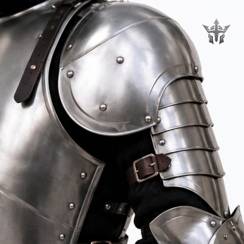 May include: A polished steel shoulder and arm armor set featuring a large rounded pauldron, articulated lames covering the upper arm, and a brown leather strap with a metal buckle. The metal plates are secured with rivets over a black garment.