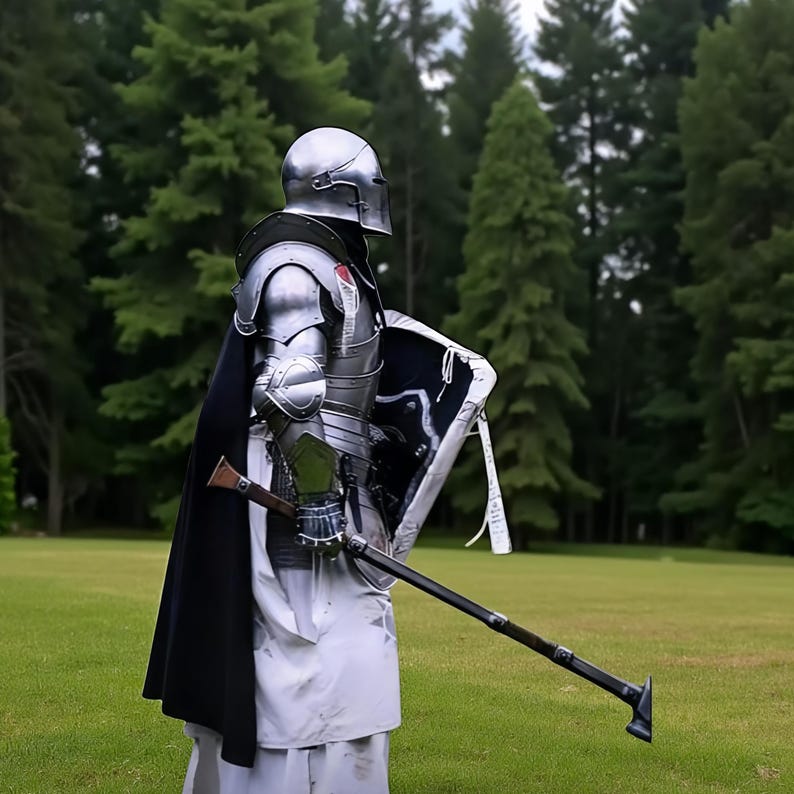 May include: A person in full silver armor, including a helmet, shoulder guards, and chest plate, stands on a grassy field. They hold a shield and a long-handled weapon. A black cape and white garment are also visible.