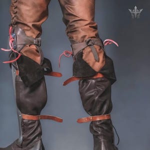 May include: A pair of dark brown leather boots featuring integrated metal greaves and knee guards. The footwear includes adjustable tan leather straps with metal buckles, black suede accents, and red cord lacing. A small trident logo is visible.