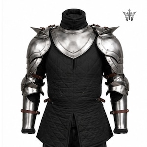 May include: A complete set of medieval armour, with polished metal shoulder and arm guards, a chest plate, and a black quilted undergarment. The armour is secured with leather straps and rivets, offering a historical aesthetic.