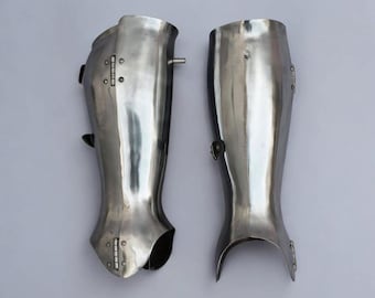 May include: A pair of polished steel greaves, part of leg armor. The metal reflects light, showing a shiny surface. Each piece has hinges and straps for attachment, and is designed to protect the lower leg. The armor is likely for historical reenactment or display.