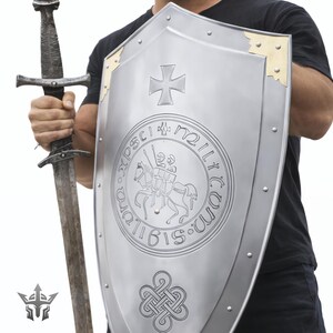 May include: A silver shield with a cross and a circular emblem, and a long sword. The shield has gold-colored corner accents and is held by a person wearing a black shirt. The sword is held in the other hand.
