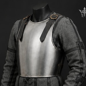 May include: A polished steel breastplate with black leather straps and buckles, worn over a grey patterned fabric garment. The armour is displayed on a black mannequin, showcasing medieval-inspired attire. The design includes decorative stitching and a belt.