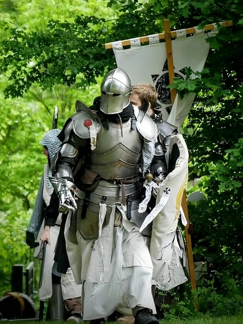 May include: A group of people in medieval armor walking through a wooded area. The armor is silver and detailed, with helmets and shoulder guards. Some wear white tunics and carry banners with symbols. The scene is set against a backdrop of green foliage.
