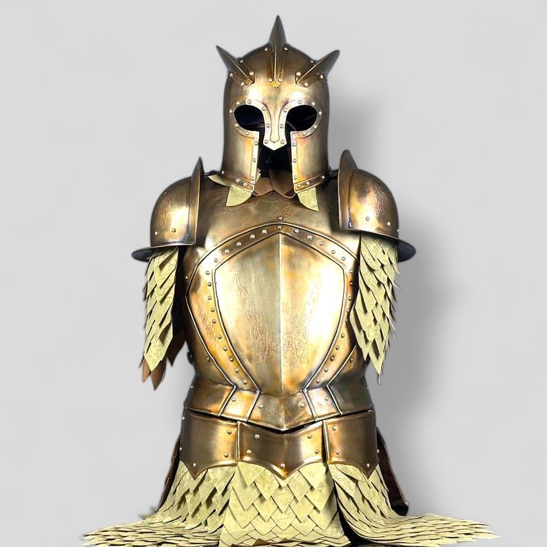 Op de afbeelding: Een goudkleurig harnas, met een helm met drie omhoog wijzende punten en ooggaten. Het harnas omvat schouderbeschermers en een borstplaat met ingewikkelde details. Het onderste deel heeft een geschubd ontwerp.