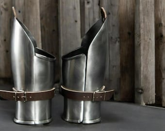 May include: A pair of silver metal arm guards with brown leather straps. The guards are designed to protect the forearm and elbow.