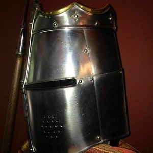 May include: A polished steel knight's helmet with gold trim and decorative details. The helmet features a visor with a horizontal opening and small ventilation holes. A spear is positioned to the left of the helmet.
