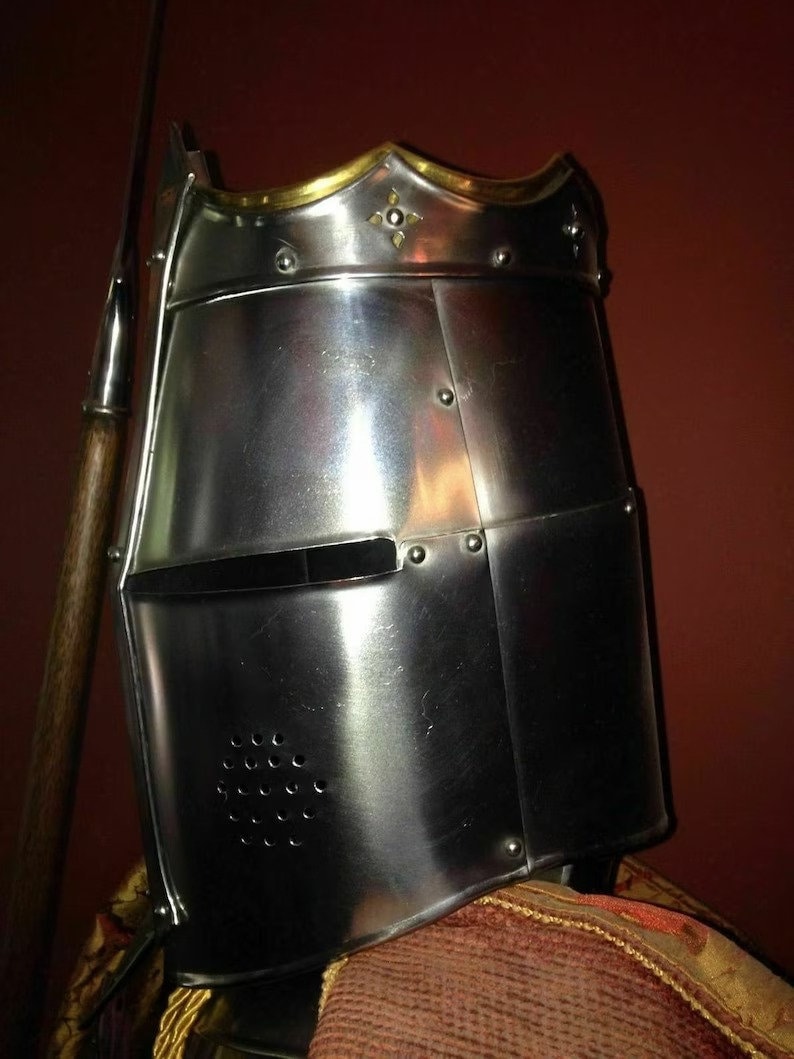 May include: A polished steel knight's helmet with gold trim and decorative details. The helmet features a visor with a horizontal opening and small ventilation holes. A spear is positioned to the left of the helmet.