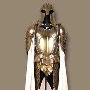 Op de afbeelding: Een volledig harnas in goudkleur, inclusief helm, borstplaat en beenbescherming. Het harnas heeft een gelaagd ontwerp en wordt gecombineerd met een witte cape. Het pak wordt tentoongesteld op een zwarte standaard tegen een bruine achtergrond.