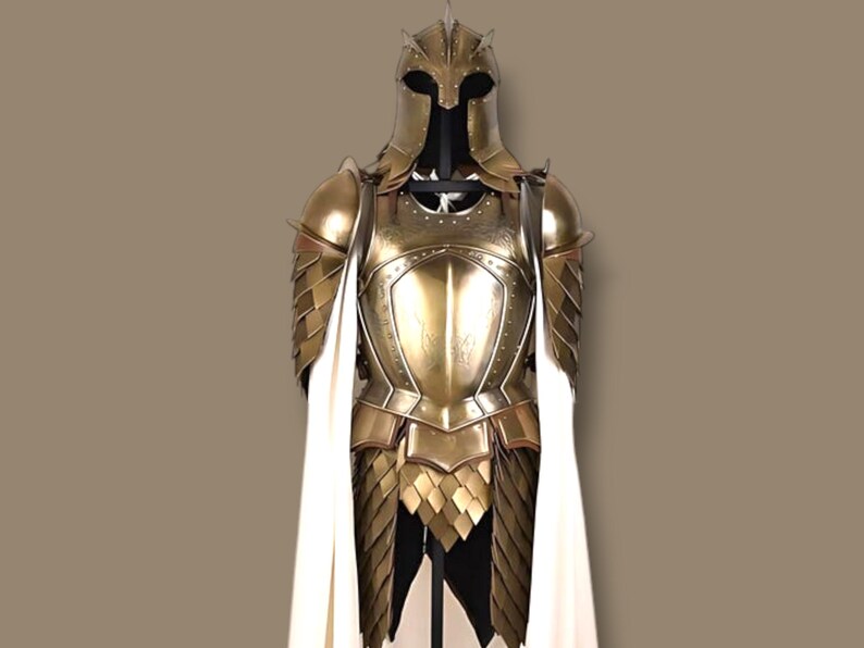 Op de afbeelding: Een volledig harnas in goudkleur, inclusief helm, borstplaat en beenbescherming. Het harnas heeft een gelaagd ontwerp en wordt gecombineerd met een witte cape. Het pak wordt tentoongesteld op een zwarte standaard tegen een bruine achtergrond.