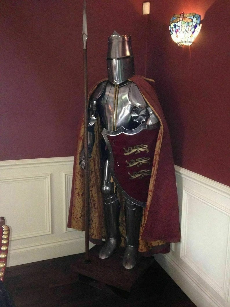 May include: A complete set of medieval armour, including a helmet, breastplate, and leg guards, stands on display. The silver armour has a red and gold cape and a shield with a crest. A spear is positioned beside the armour.
