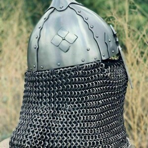 May include: A medieval-style helmet with a pointed top and a decorative floral emblem. The helmet is made of shiny metal and is riveted together. A chainmail neck guard is attached to the helmet.