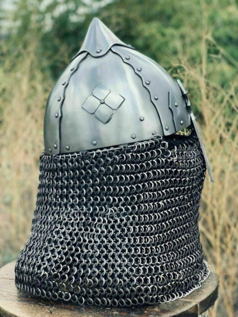 May include: A medieval-style helmet with a pointed top and a decorative floral emblem. The helmet is made of shiny metal and is riveted together. A chainmail neck guard is attached to the helmet.
