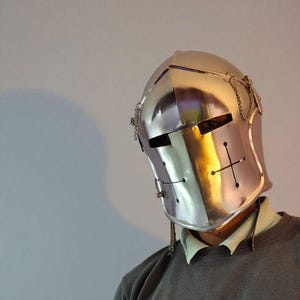 May include: A polished silver knight's helmet with a cross design and chain accents. The helmet has eye slits and a hinged visor. The metal reflects light, creating a bright, shiny appearance. The helmet is a historical item.