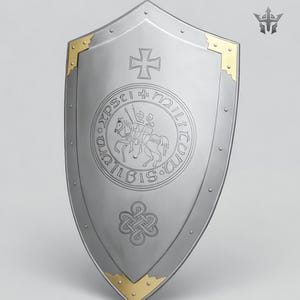 May include: A silver-colored shield with a pointed bottom and rounded top. The shield features a cross at the top, a circular emblem with a knight on horseback, and gold-colored corner accents. The shield has a medieval theme.