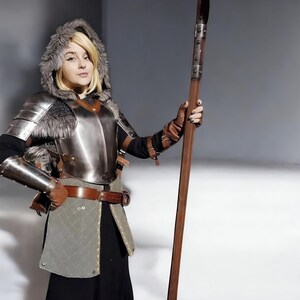 May include: A person in a medieval costume, featuring a metal breastplate, fur-trimmed hood, and brown leather gloves, holding a long wooden spear. The outfit includes a quilted skirt and a brown leather belt. The costume is reminiscent of historical armour.