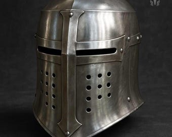 May include: A silver-colored medieval helmet with a cross design on the front. The helmet has a visor with eye slits and numerous small ventilation holes. The helmet is made of metal and has a slightly aged appearance.