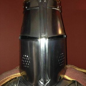 May include: A polished steel knight's helmet with a pointed top and riveted construction. The helmet features a visor with eye slits and ventilation holes. A portion of the knight's armour and a brown and gold cape are visible.