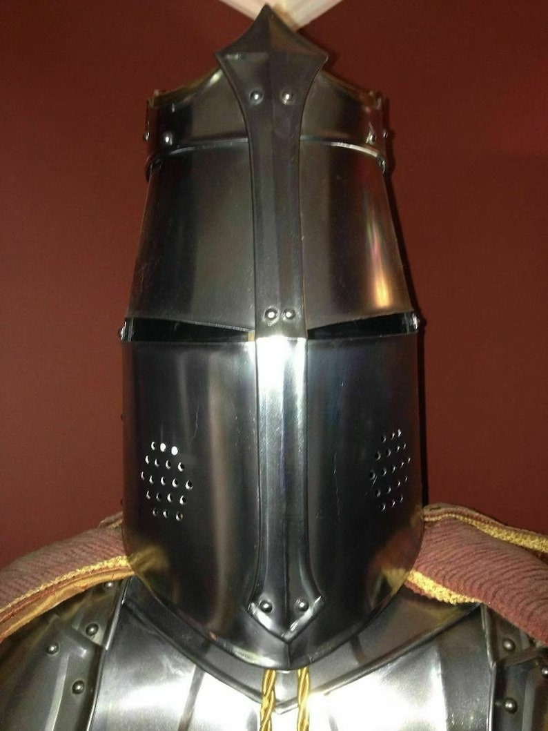 May include: A polished steel knight's helmet with a pointed top and riveted construction. The helmet features a visor with eye slits and ventilation holes. A portion of the knight's armour and a brown and gold cape are visible.