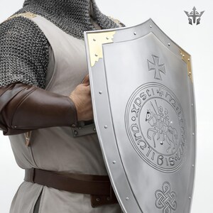 May include: A silver shield with a cross and a circular emblem featuring a knight on horseback. The shield has gold corner accents and is riveted around the edges. The shield is held by a person wearing chainmail and brown leather arm protection.