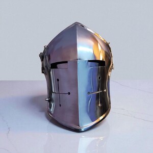 May include: A polished, silver-colored medieval helmet with a cross design on the front. The helmet has a smooth, reflective surface and a visor with eye slits. The helmet is designed to protect the head and face.