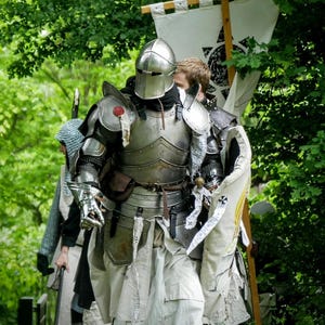 May include: A suit of polished steel plate armor featuring a closed helmet, pauldrons, and gauntlets, worn over a white tunic. A shield with a cross emblem is held at the side. The text Middle Age Medieval is displayed at the top of the image.